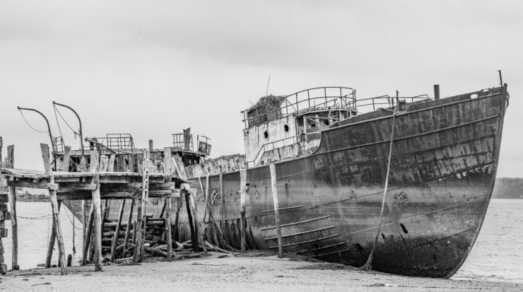 A pier leads to a rusting ship sitting on the mud at low tide.