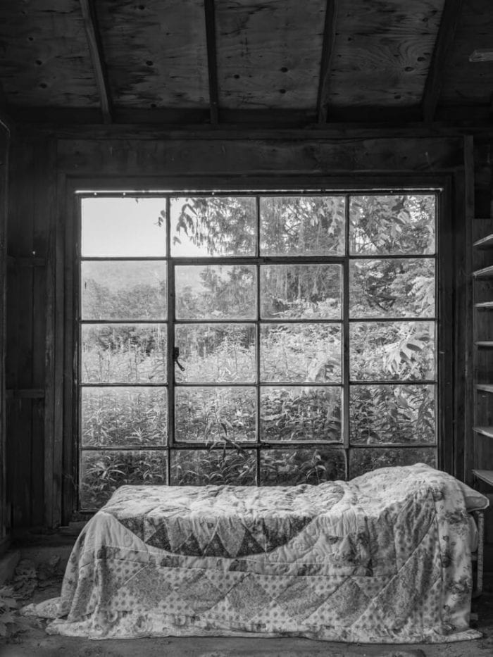 A bed with a quilt lies forgotten in a barn with a large window above it