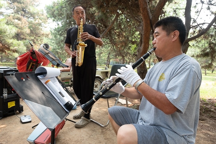 A man playing the saxophone and another man playing a clarinet play in the park.