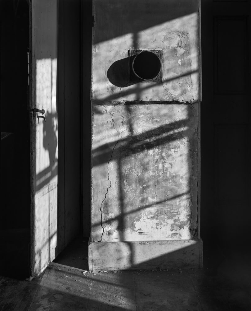 Where stove once stood in the​ kitchen of the Olson House. The open door leads to the​ barn and the stove pipe is lit by afternoon​ sun.